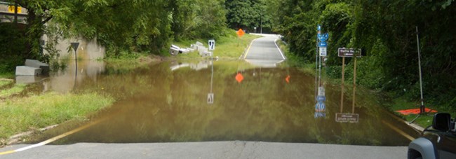 A fifty foot wide dip in the road at Kittatinny Point, NJ is filled with flood waters from heavy rains. Road sings can be seen past the water filled dip.