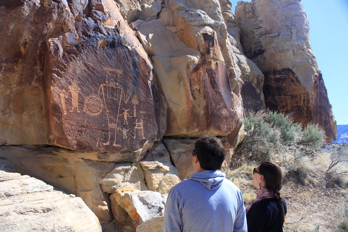 Visitors view an iconic petroglyph panel at McKee Spring, featuring several humanoid figures. The large central figure appears to hold a bell-shaped object in one hand, and a circular shield or sun with a swirling design.