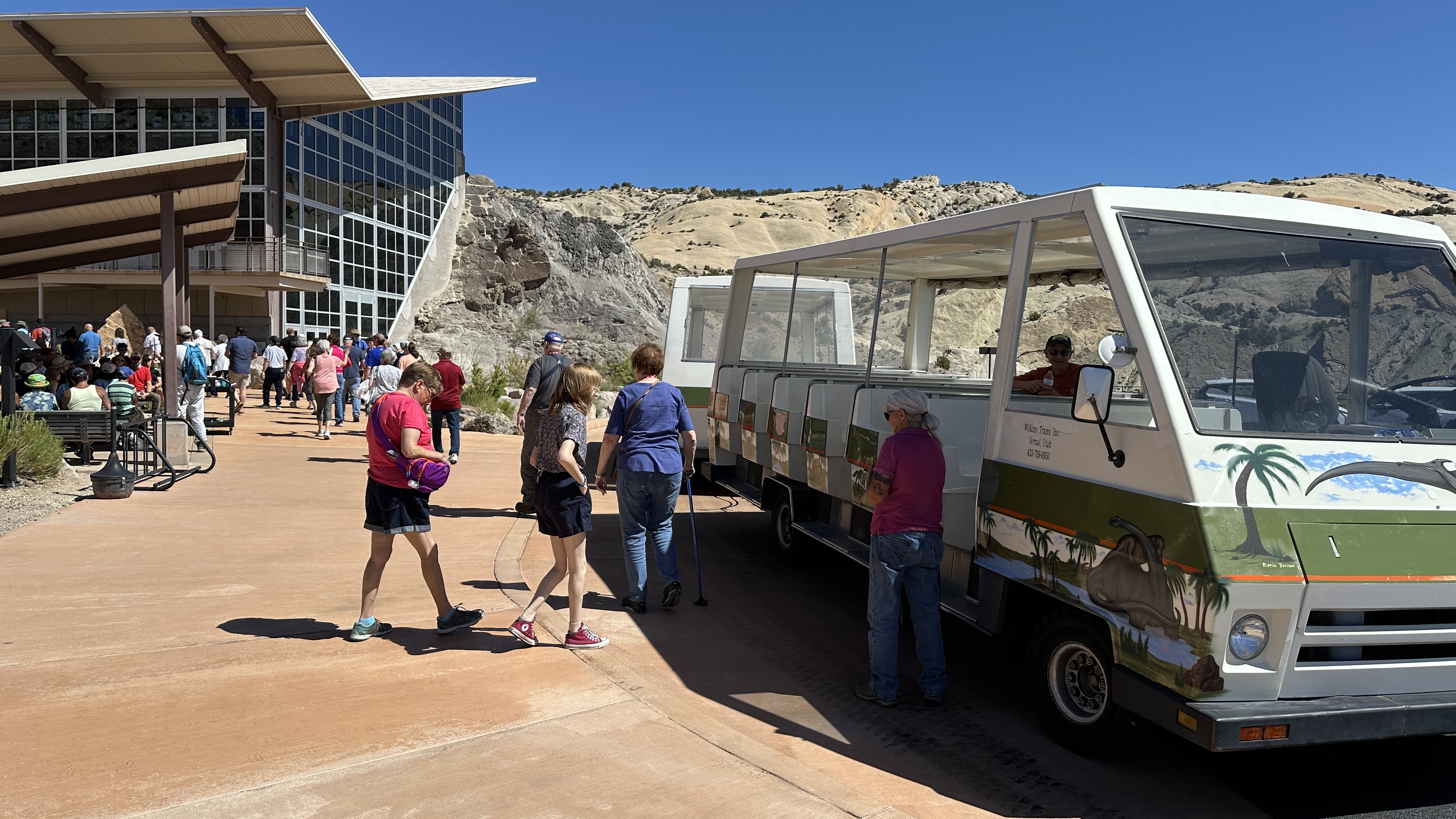 An open air shuttle sits in a parking lot as peopl walk towards a building with a slanted roof.