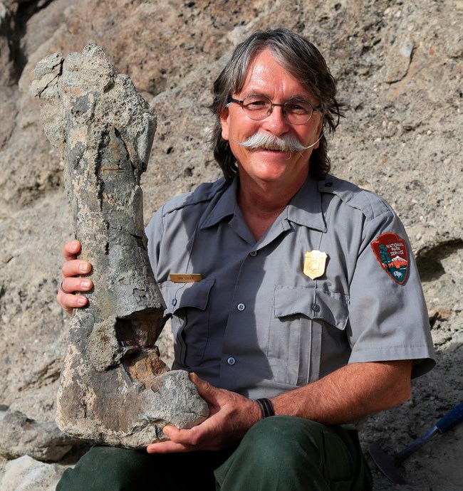 A photo of a man with a fun mustache smiling while sitting on a rock, holding a fossil.