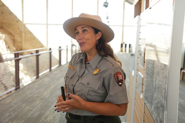 A woman with brown hair and a park service uniform explains something.