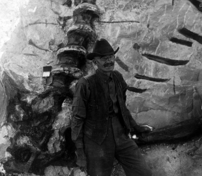 A black and white photo of a smiling man standing in front of a rock face with dinosaur fossils exposed.