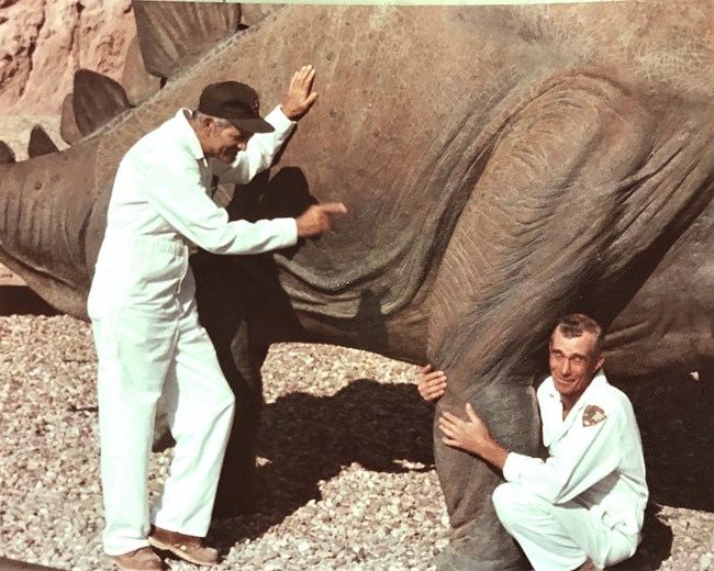 Two men in white jumpsuits are smiling beside a stegosaurus statue. The left one points at the dinosaur while the right one hugs its leg.