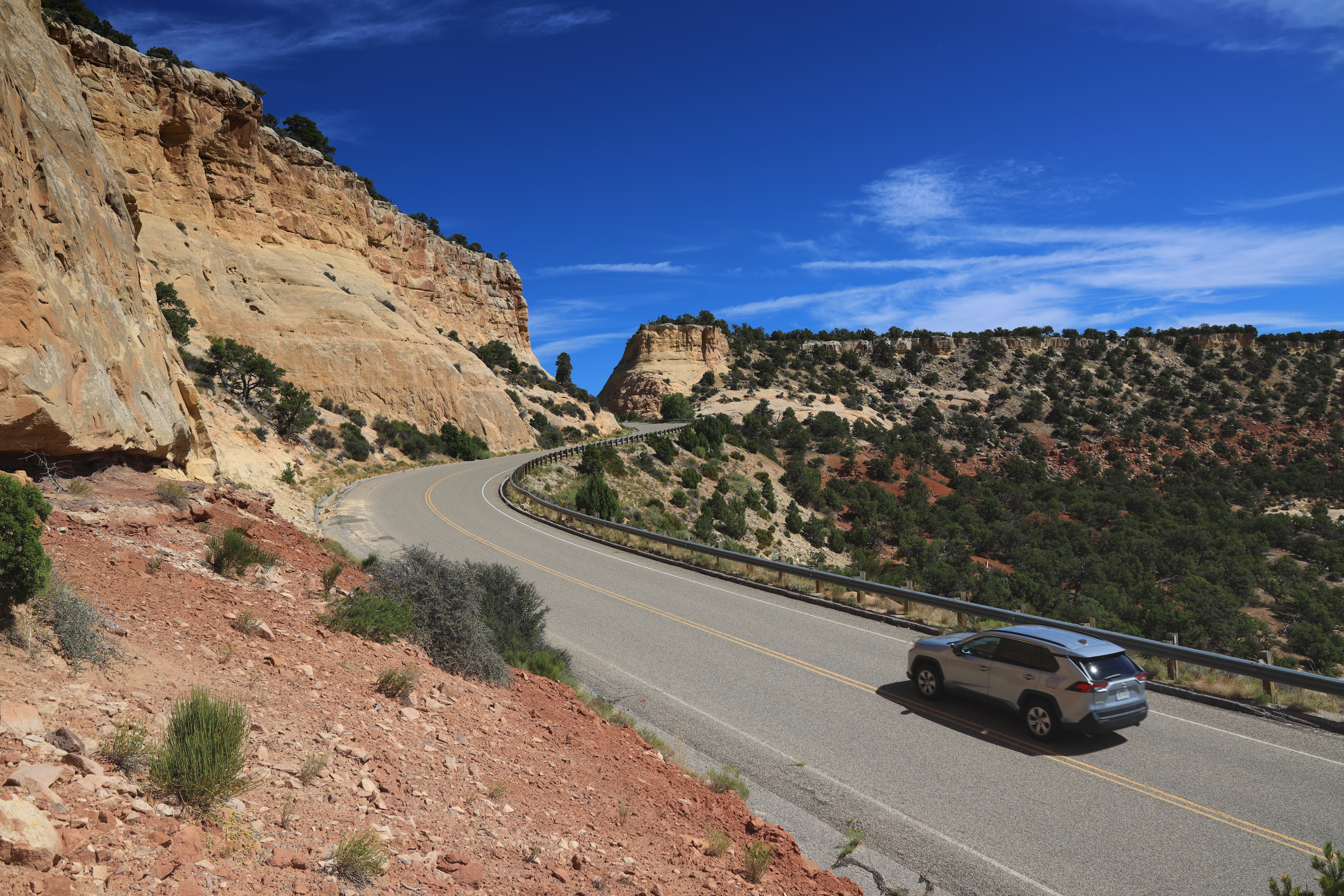 A silver vehicle drives on a road through color cliffs.
