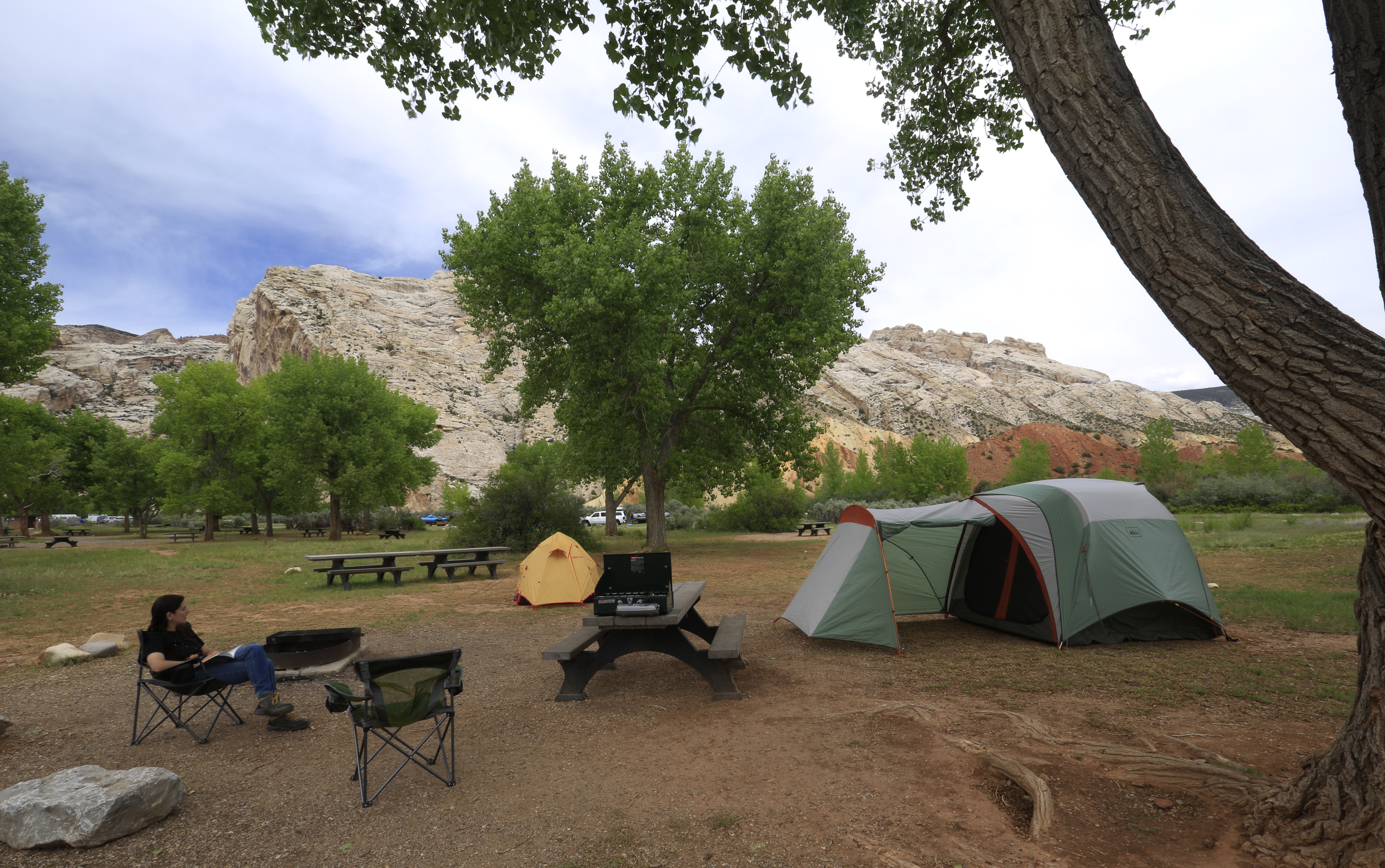 A woman sits in a folding chair near a picnic table and tent. Trees and a rocky mountain fill the background