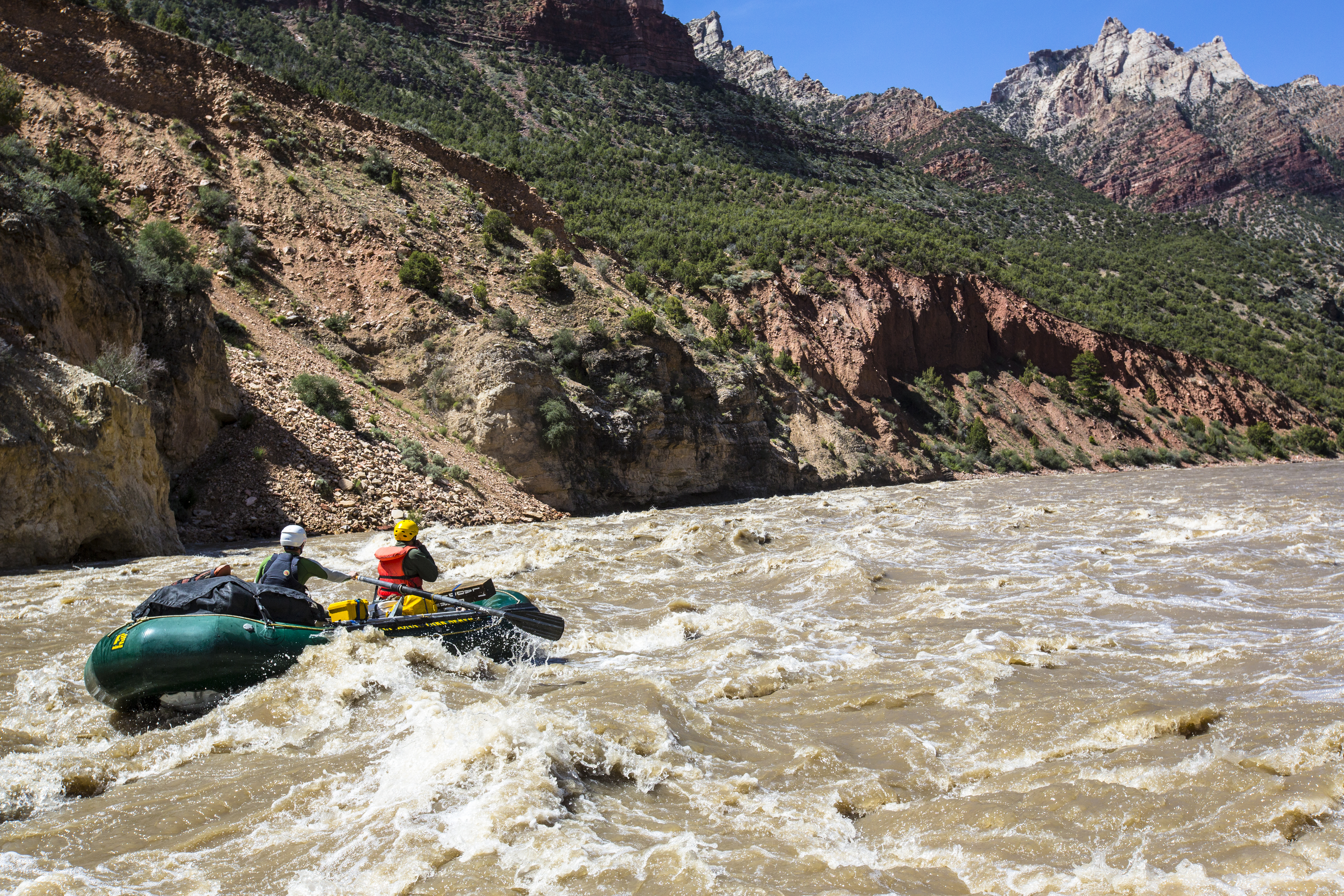 A green raft float through a white water rapid as mountains rise in the background.