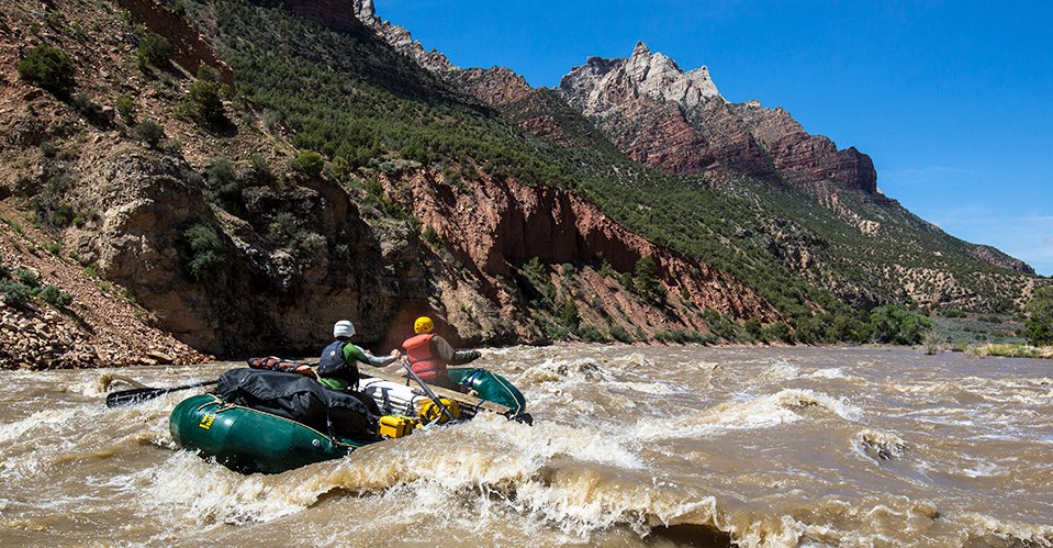 2 people ride a green rubber raft as it runs through a white water rapid. In the distance a tall mountain rises into a blue sky.