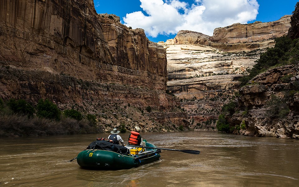 2 people in a green rubber raft float on a calm river as cliffs of yellowish tan rock tower above them