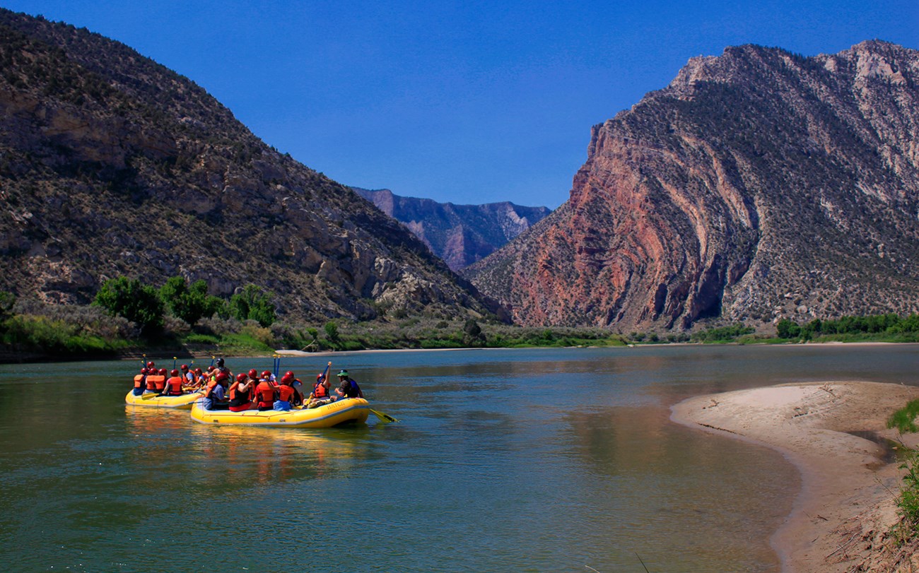 Two yellow rafts filled with people float on a rivers towards large colorful cliffs.
