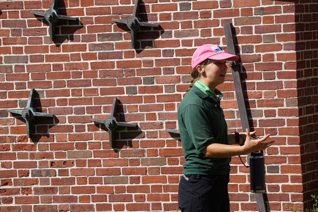 A woman standing in front of a brick wall with metal trivets, holding her hands out and speaking