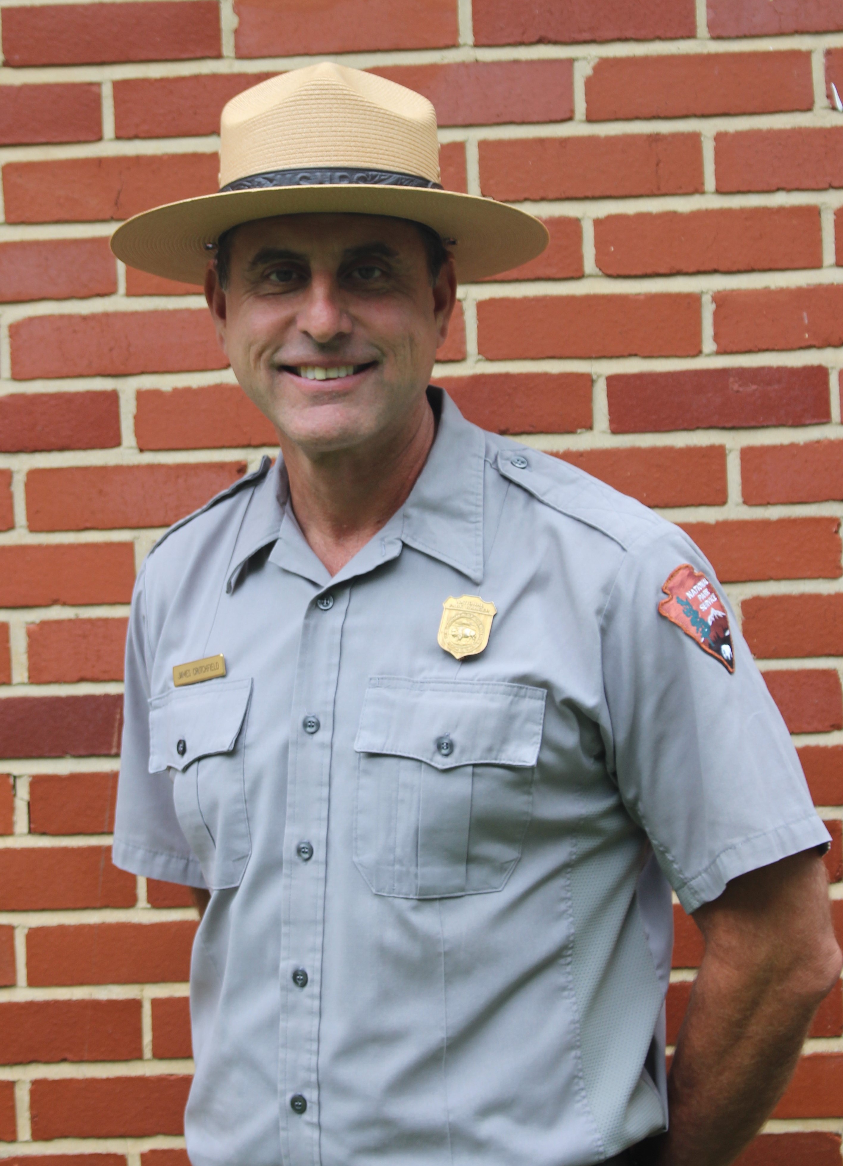 A smiling man in a park ranger uniform and classic flat hat stands in front of a brick wall.