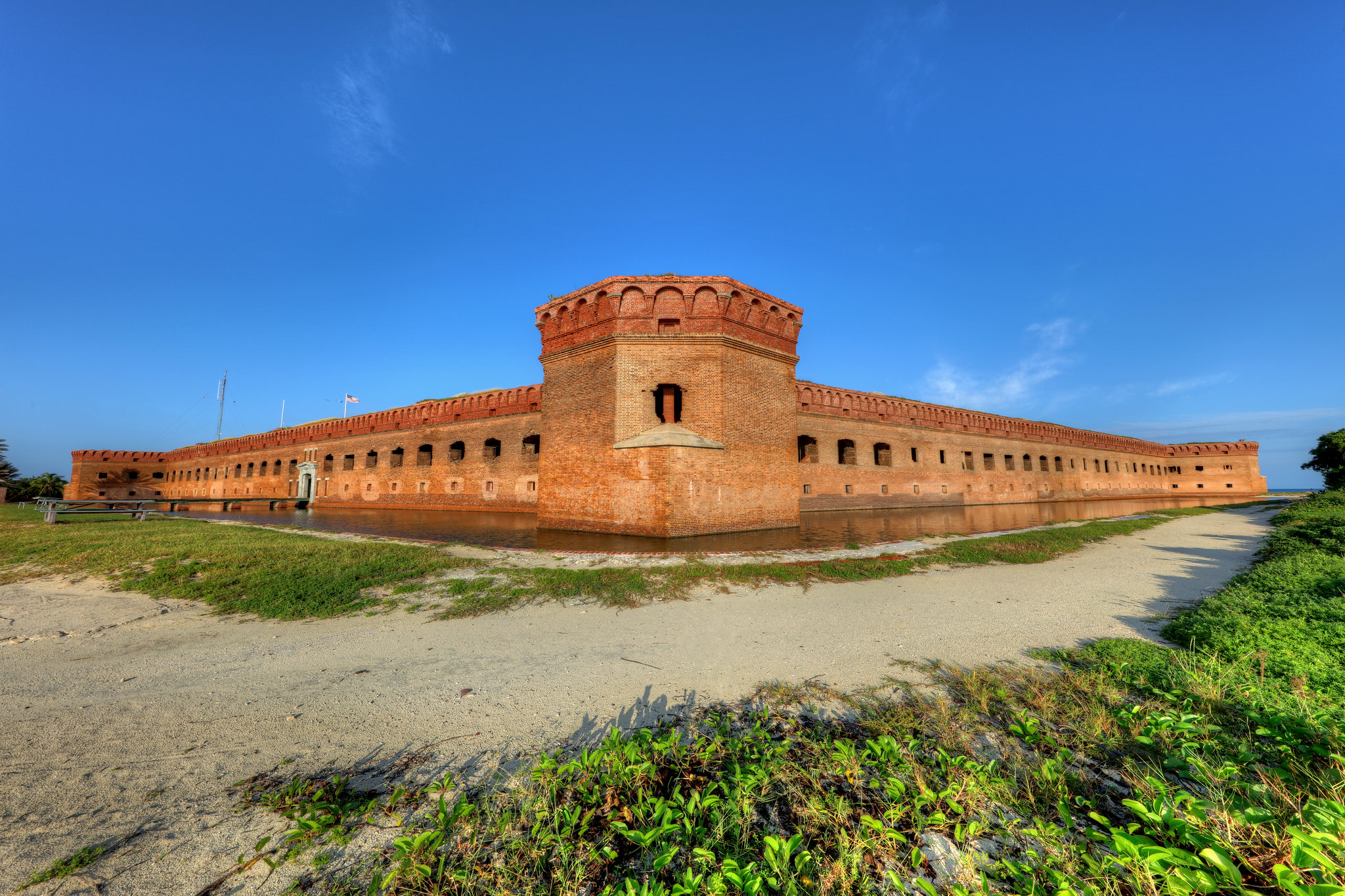 NPS Photo - Fort Jefferson on Garden Key in Dry Tortugas National Park A landscape photo of Fort Jefferson's corner bastion and brick walls. The sky is clear blue and green grass and sand are visible in the foreground.
