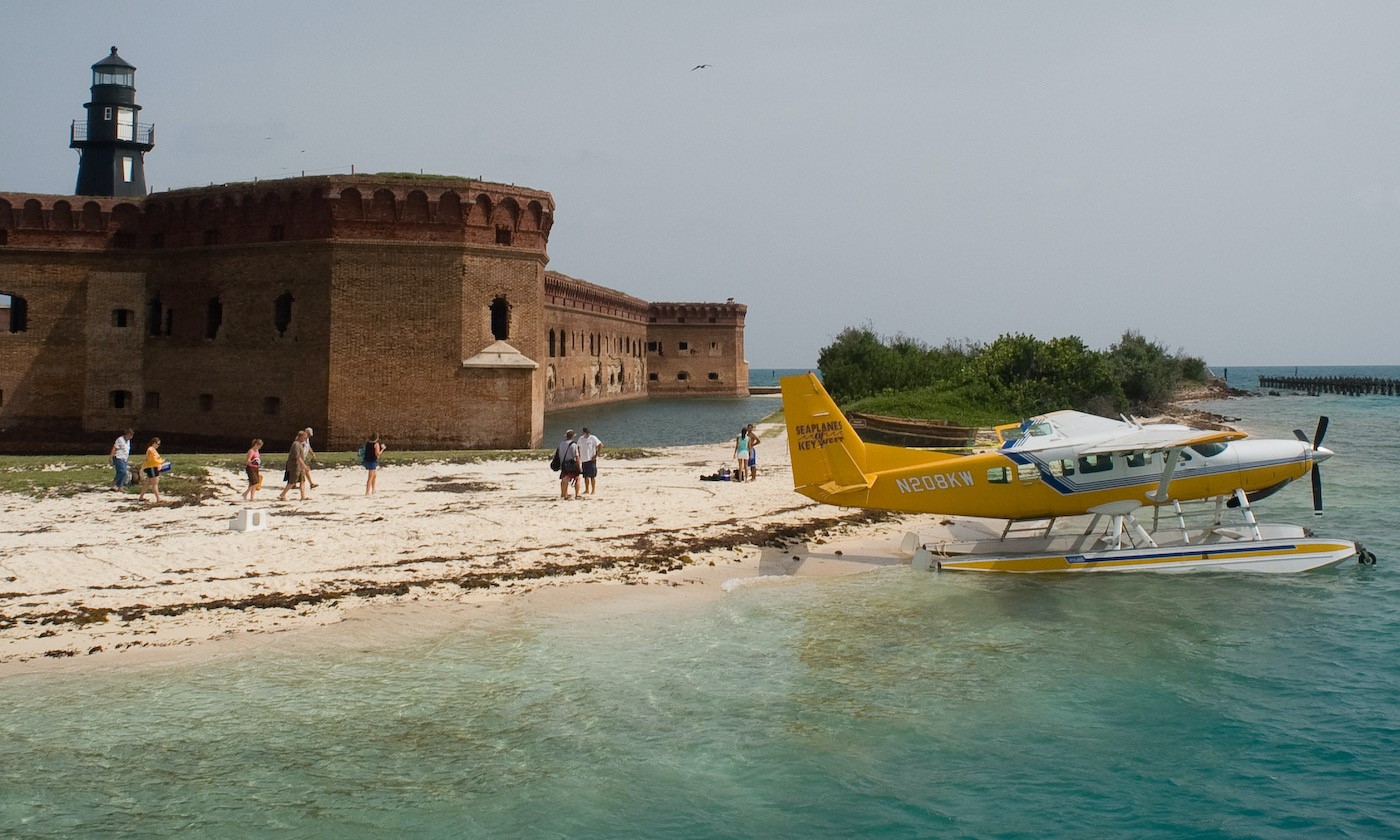 Sea_plane_at_Dry_Tortugas_National_Park A sea plane sits on the beach as visitors walk toward it with three-story brick fort with harbor lighthouse in the background. Blue-green water in the foreground.