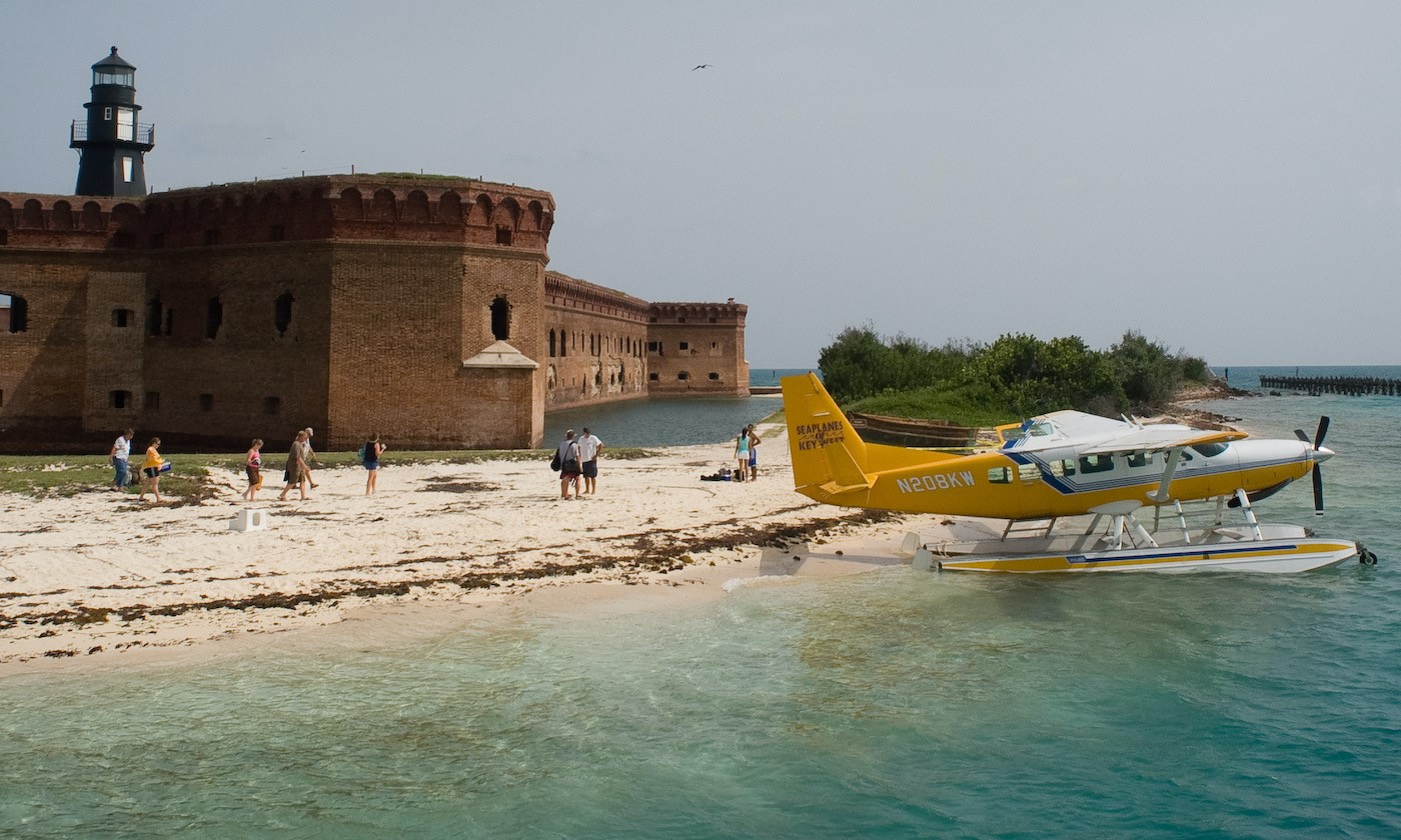 A sea plane sits on the beach as visitors walk toward it with three-story brick fort with harbor lighthouse in the background. Blue-green water in the foreground.