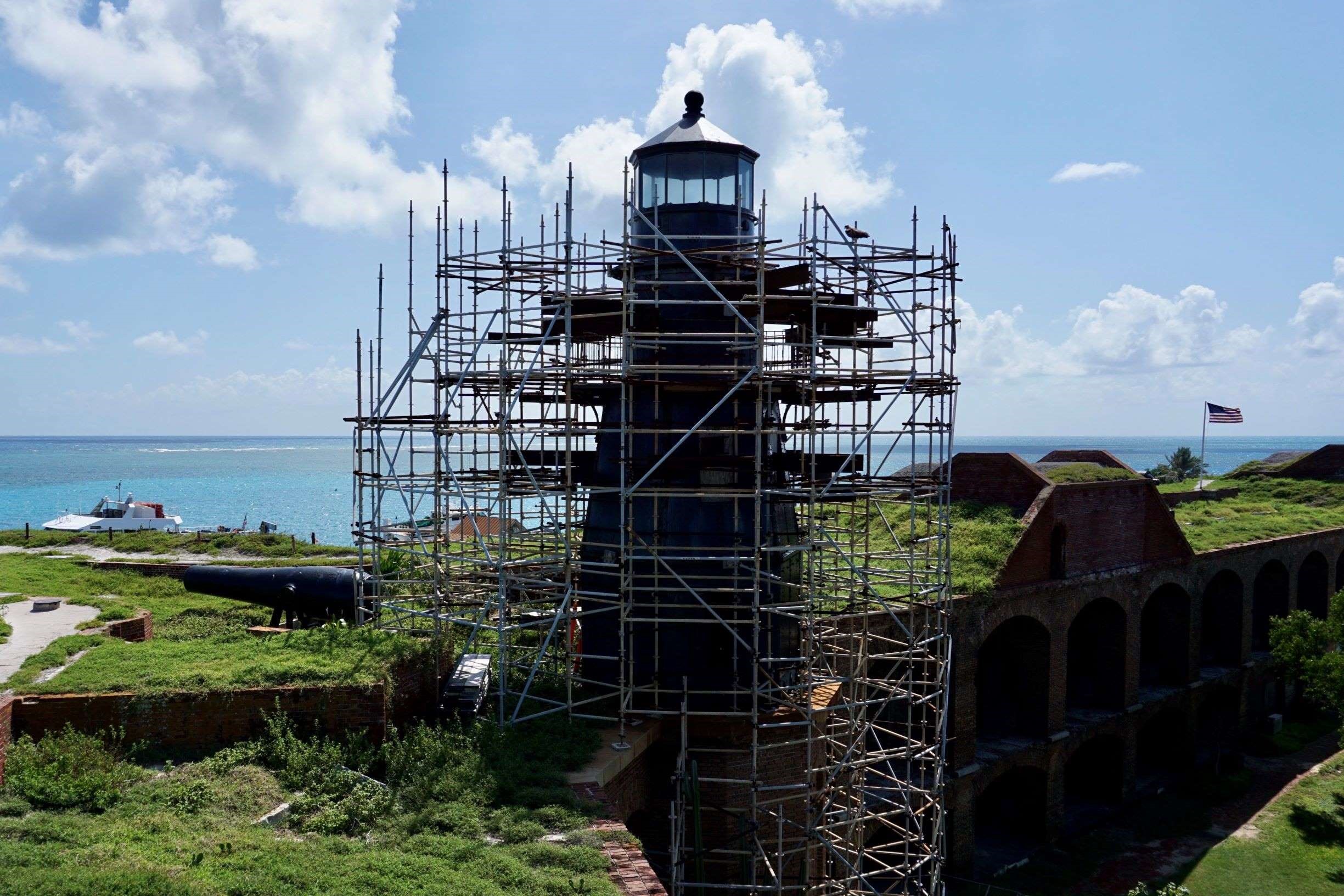 Lighthouse encased in scaffolding sits atop third level of brick fort with ocean behind.