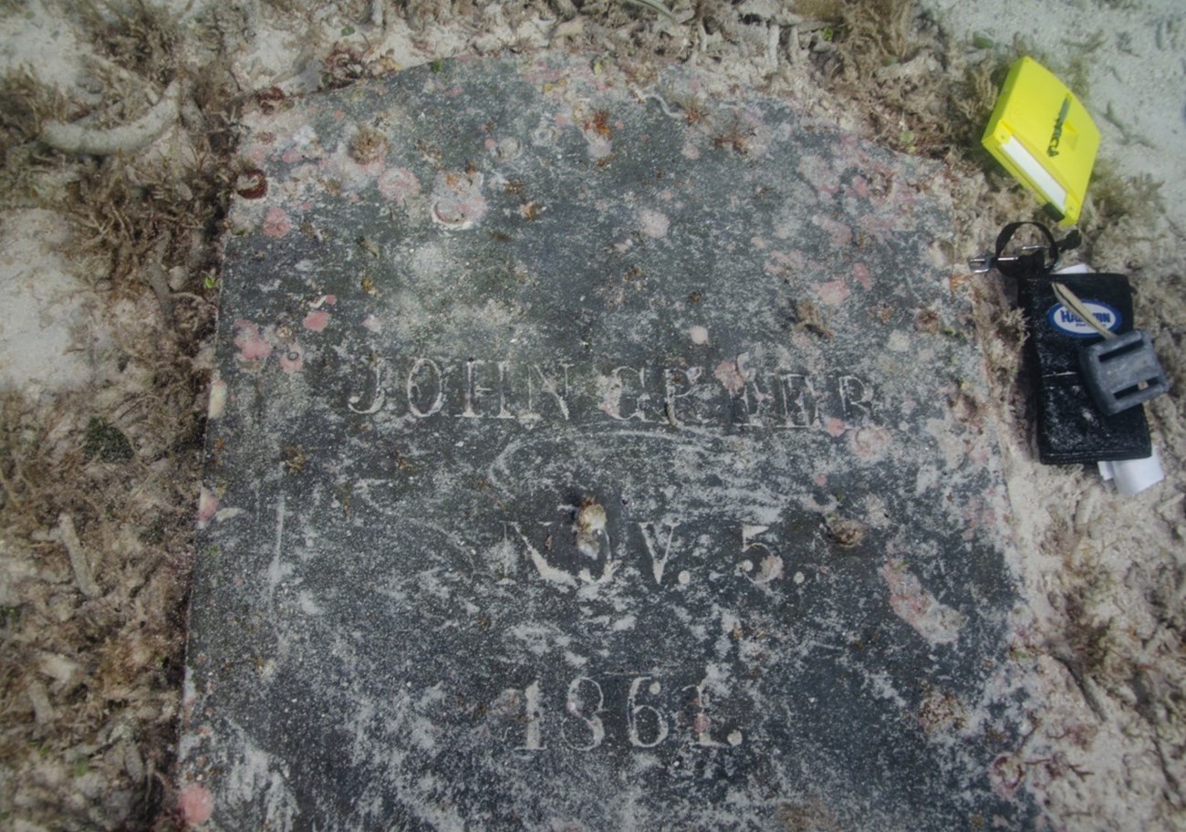 A gravestone with sand shows the inscription "John Greer. Nov. 5. 1861."