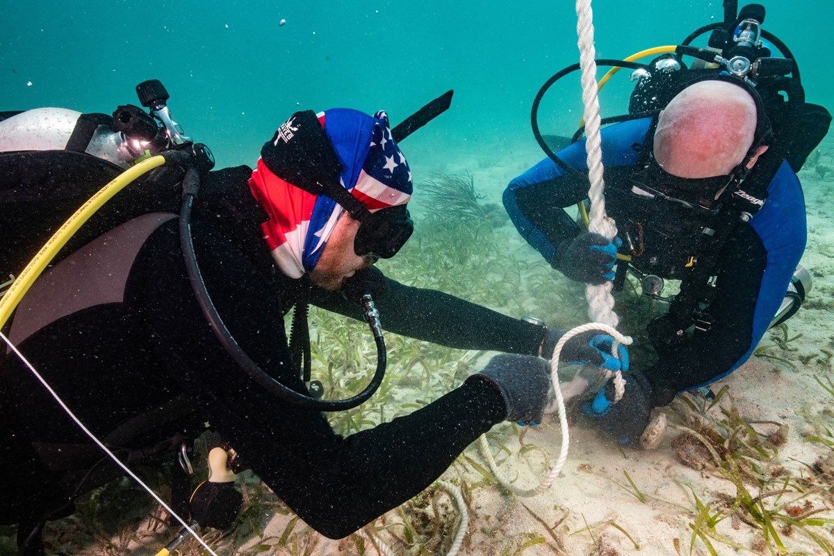 Two divers underwater secure a line of rope to the sandy ocean floor