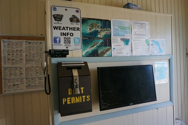 A wall covered in signs, maps, a radio, a screen, and a wooden box labeled "BOAT PERMITS"