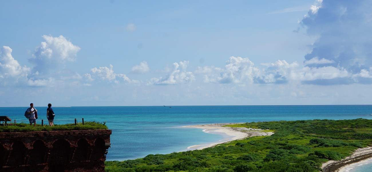 An island, blue ocean, and blue sky with vegetation being overlooked by 2 people standing on a brick structure.