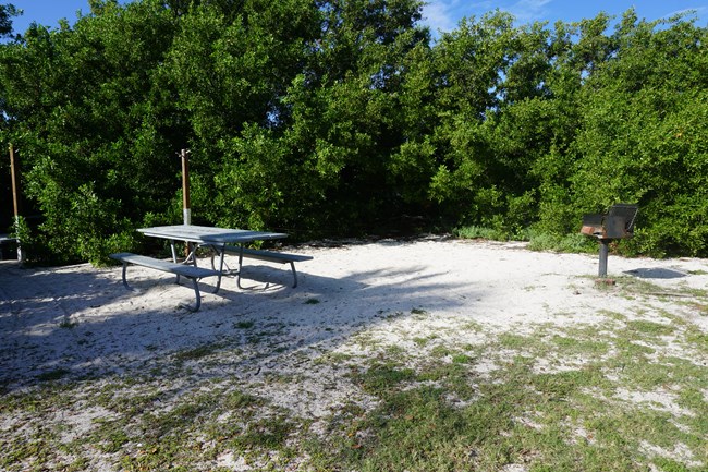 A picnic table and metal grill on a sandy and grassy area, surrounded by shrubbery