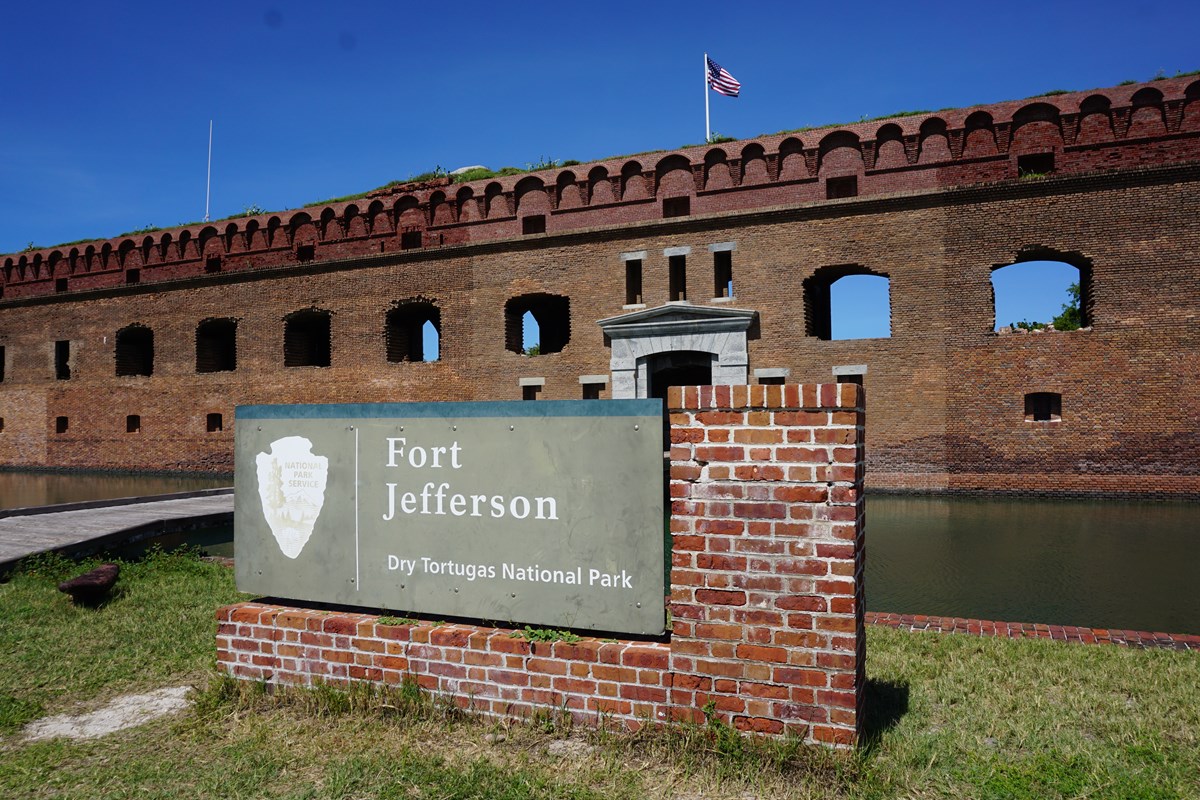 A sign in front of a large brick structure, showing the National Park Service arrowhead and the words "Fort Jefferson Dry Tortugas National Park"