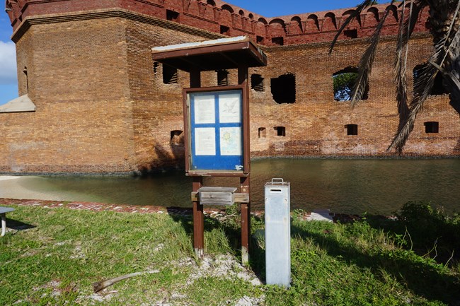 A wooden sign and a metal box on grass with a brick structure in the background