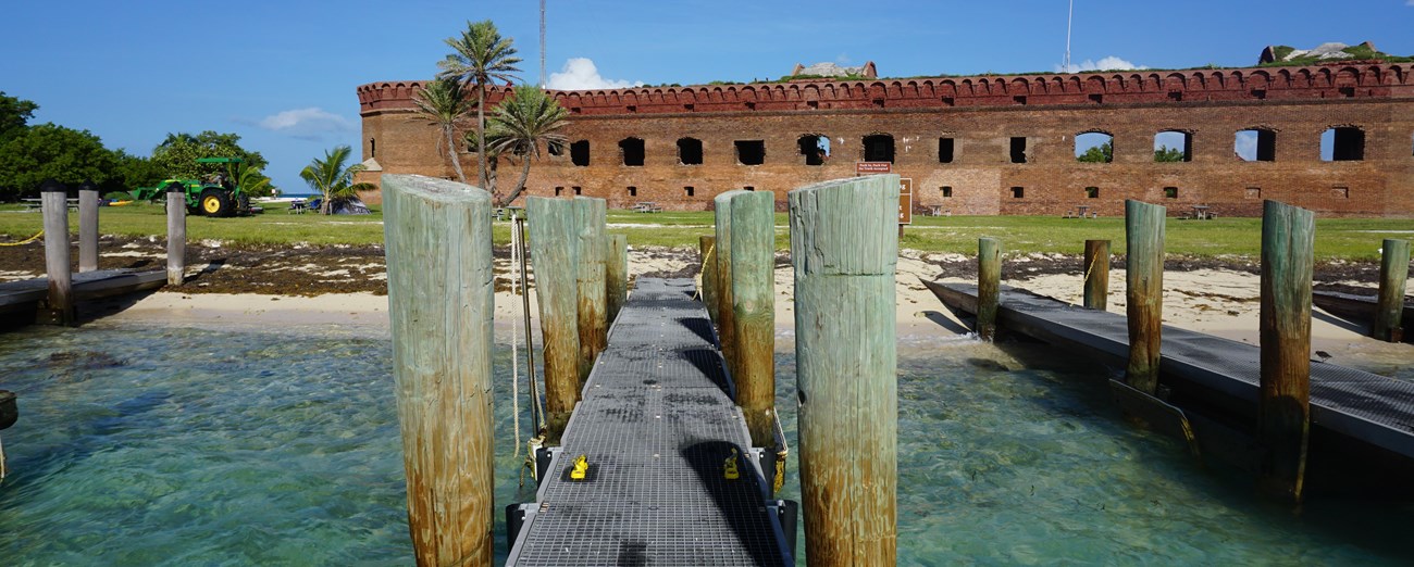 A wooden pier around sand and blue ocean waters, with a large brick structure in the background