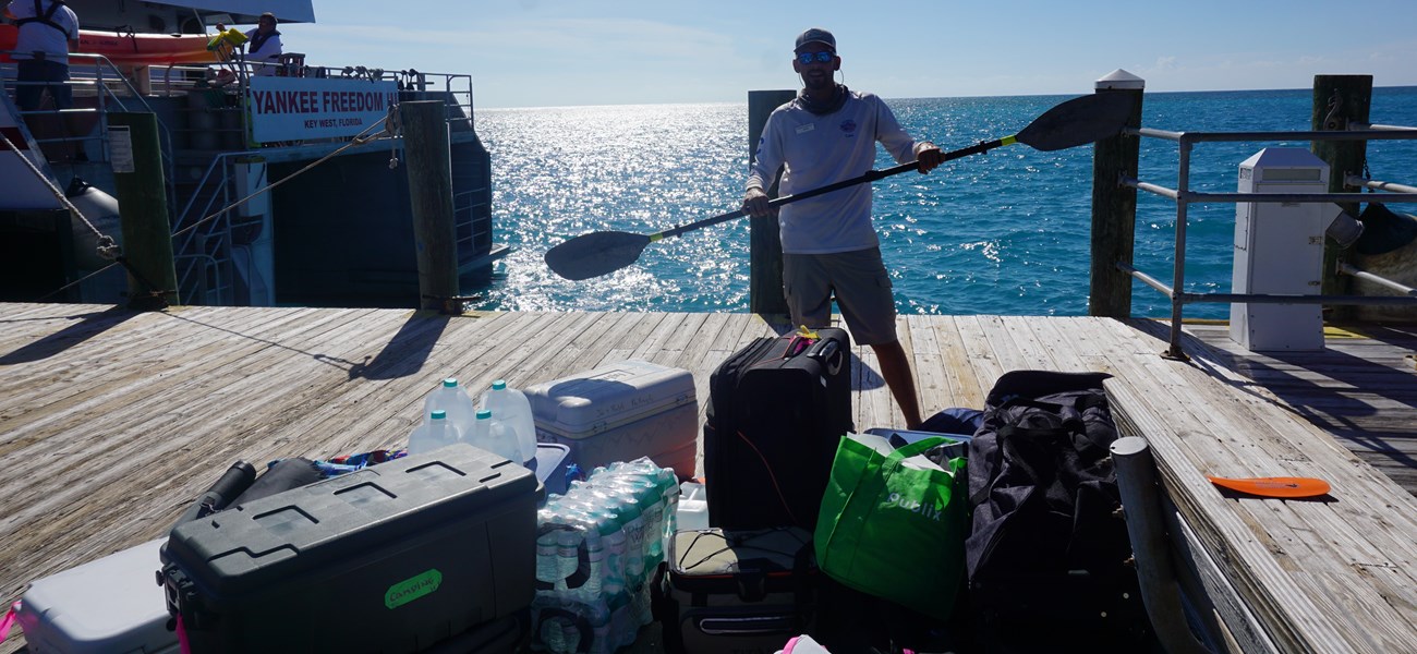 A man holding a kayak paddle, standing on a dock beside camping gear and a boat