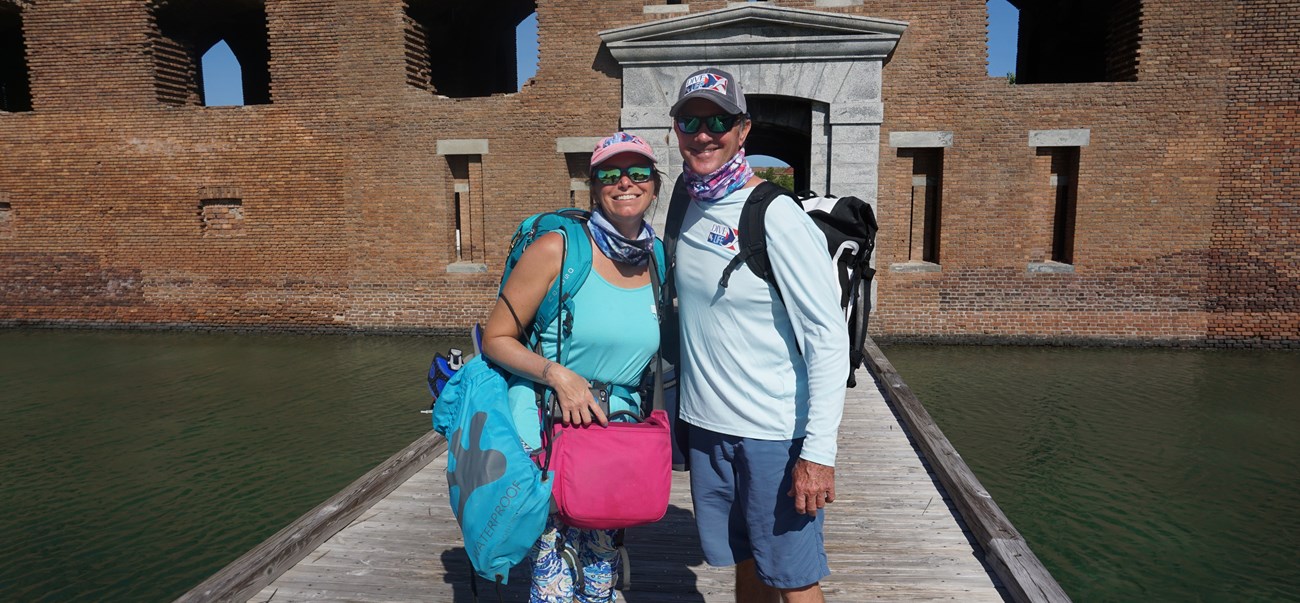 2 people smiling and standing on a wooden boardwalk leading to a large brick Fort