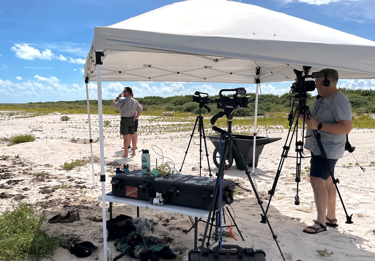 A white tent with film equipment is set up on the beach with a park ranger in the background.