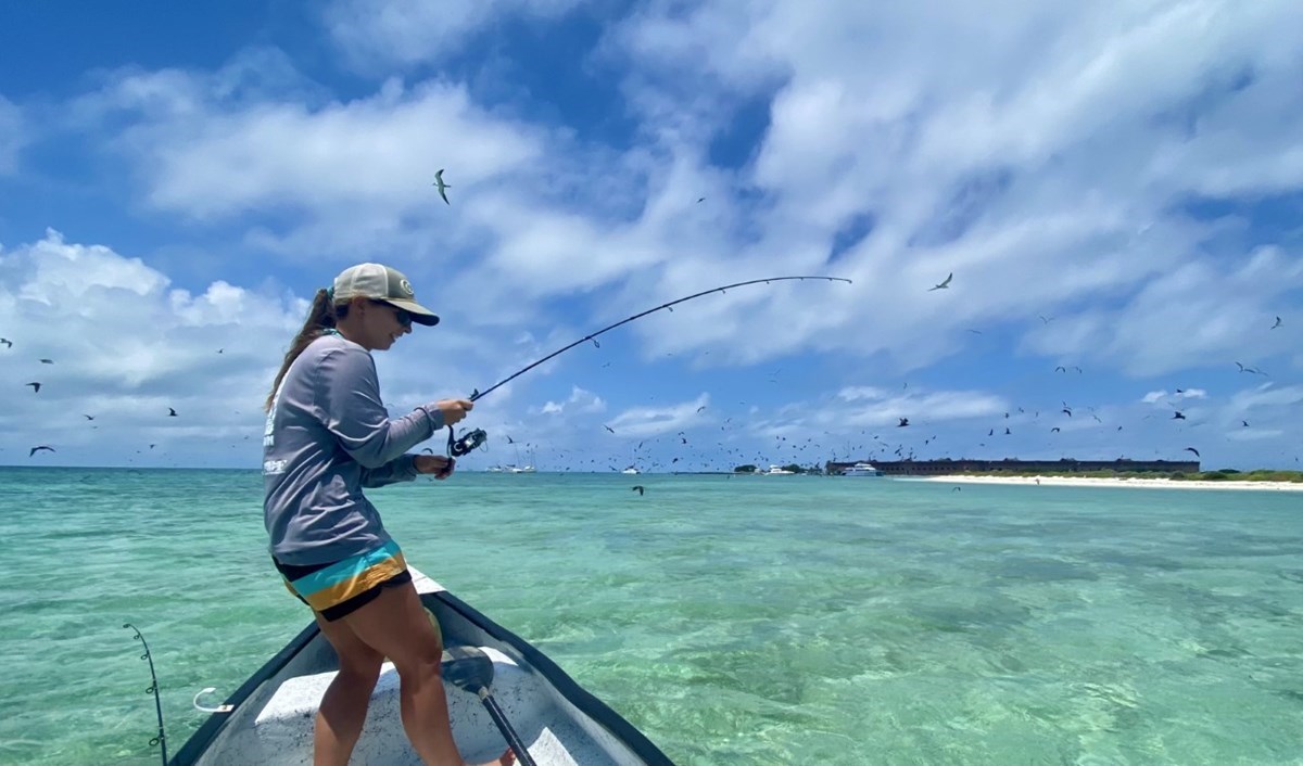 A person holding a bent fishing rod stands on a boat in clear, turquoise waters.