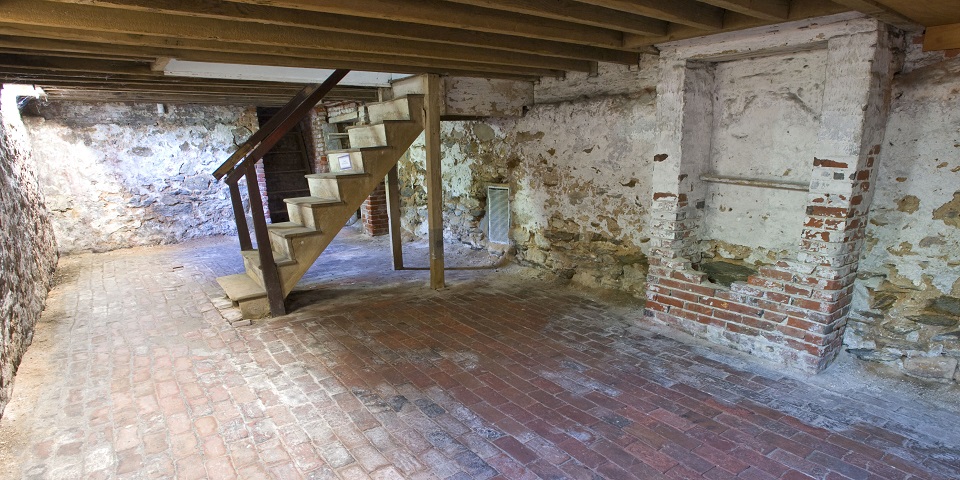 Color photo of an empty basement with brick floors, stone walls, and a wooden staircase descending from the wooden ceiling. Color photo of an empty basement with brick floors, stone walls, and a wooden staircase descending from the wooden ceiling.