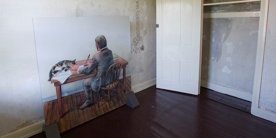 Color photo of a large illustration mounted on a wall that shows a man sitting at a desk with his back to the viewer while a calico cat reclines on the desk next to a piece of paper.