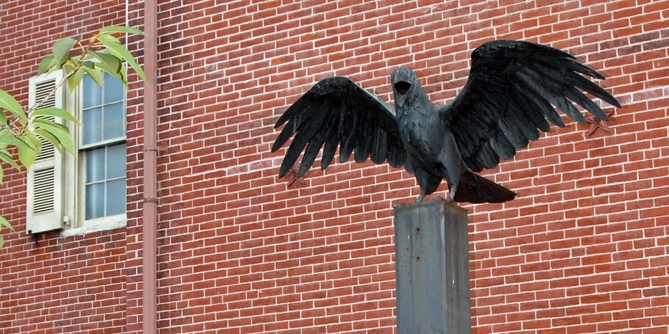 Raven outside Poe House Color photo showing a large raven statue with wings outspread on a metal plinth in front of a red brick wall.