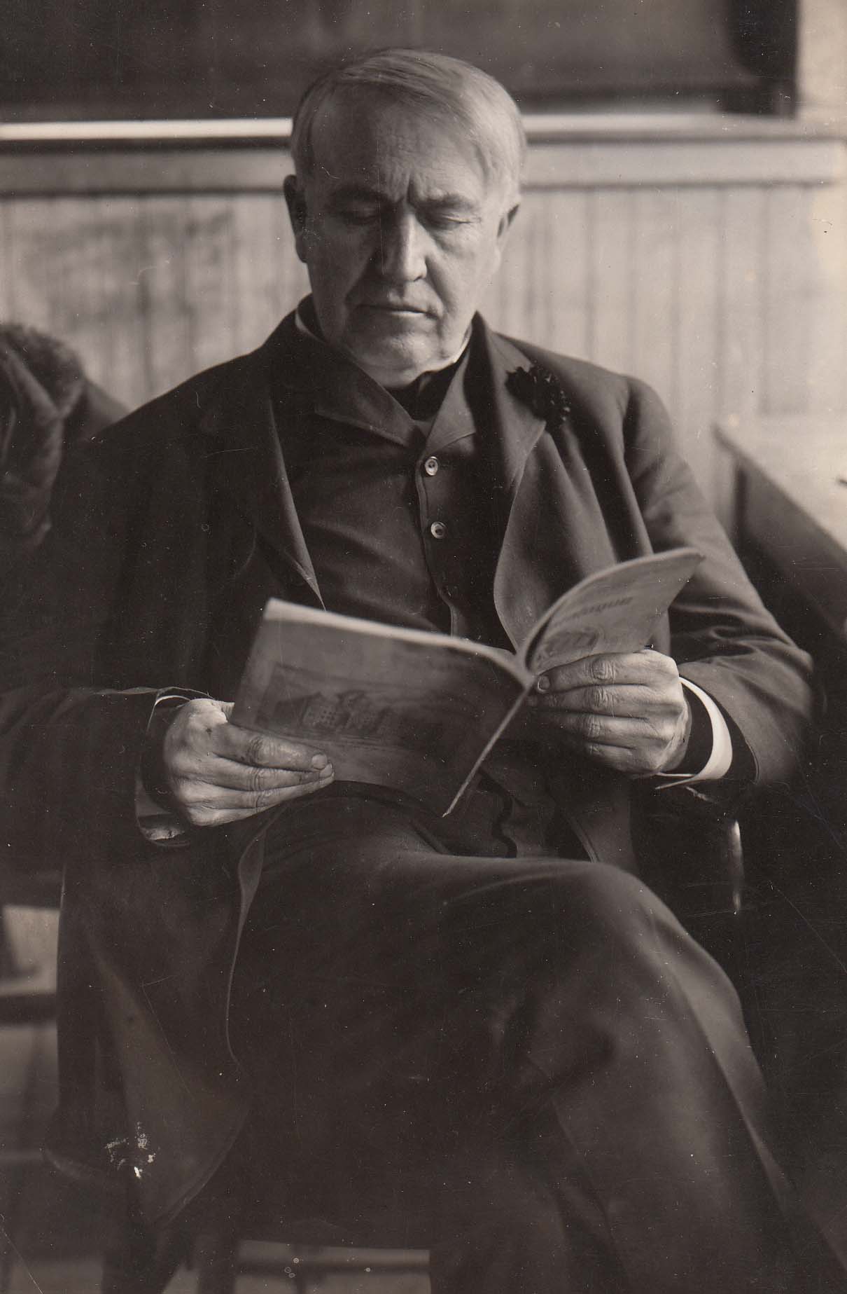 Thomas Edison, in a dark three piece suit, is sitting in a chair reading a book.