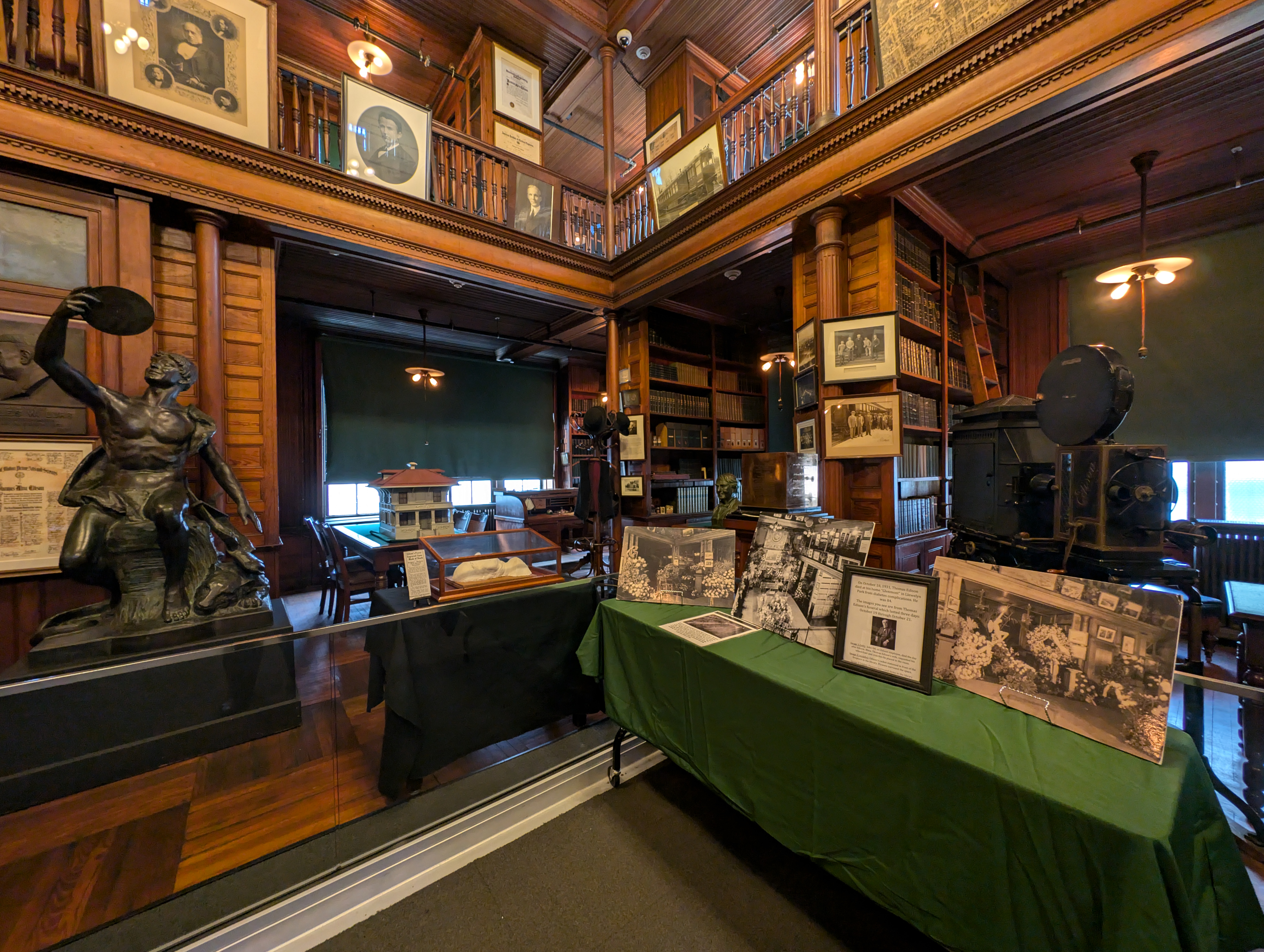Edison's Library in October 2024 Edison’s library with wood paneling, portraits on a mezzanine, and a table displaying black-and-white photos from his funeral, including flower arrangements around his coffin. A bronze statue is on the left, and a vintage projector is on the right.