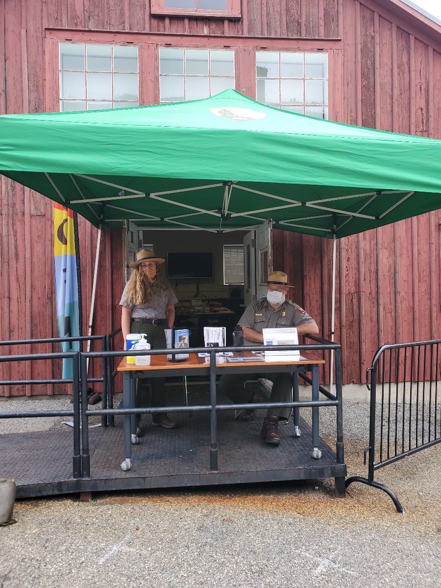 Large green tent in front of a red barn like building.  Two park rangers are under the tent.