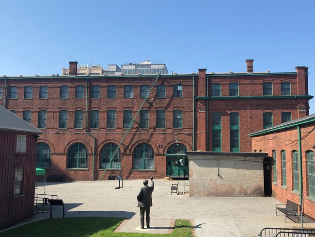 Brick, wood and concrete buildings, statue and gravel courtyard. Sky in background.