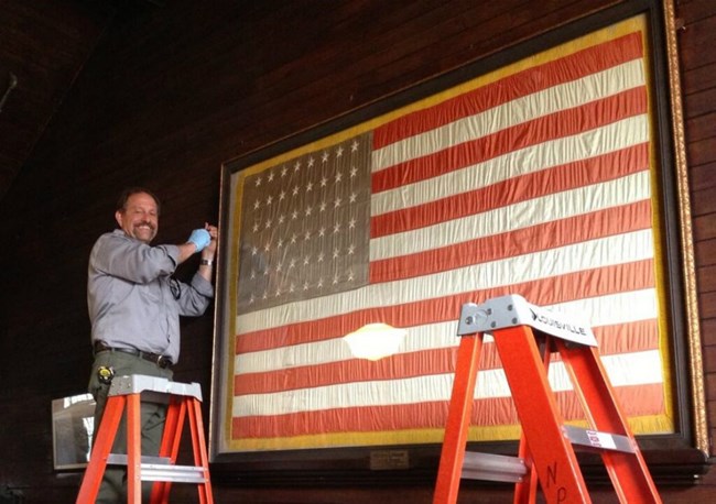Ranger standing on chair re-installing a newly conserved 48 star flag in the main laboratory entrance hall.