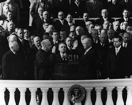 Black and White Photograph of Dwight Eisenhower raising his right hand, taking the Oath of Office. Vice President Richard Nixon is pictured on far right; former president Harry Truman on far left. 