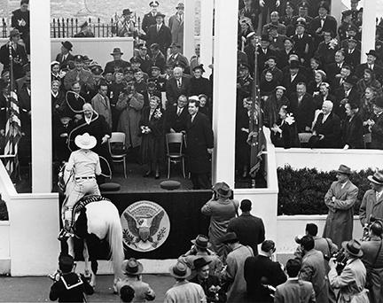 Black and White Photograph of Marty Montana, on a white horse, lassooing a laughing President Dwight Eisenhower while standing on the reviewing stand. Mamie Eisenhower and Richard Nixon are both in the photograph, laughing. 