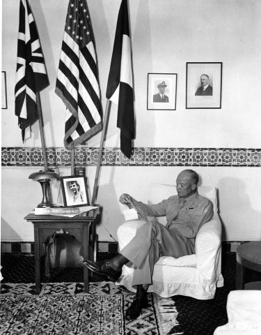 Dwight Eisenhower sits on a white chair, reading a newspaper, while behind him are three flags of Allied powers, including the United States flag. On a table next to the chair is a framed photograph of Mamie Eisenhower.
