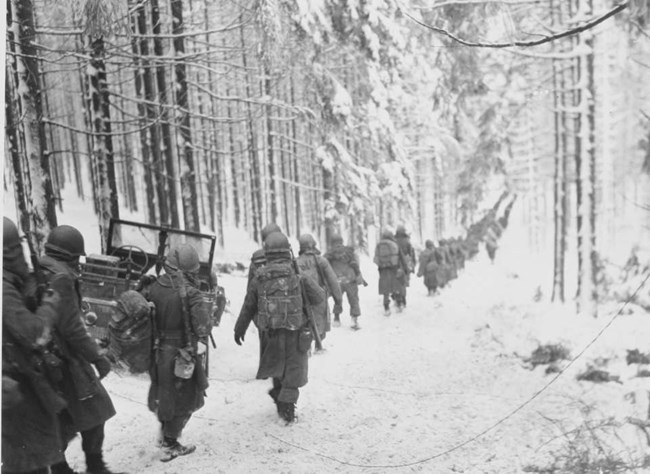 Black and White Photograph Showing American Soldiers Walking down a snow covered road in a snow covered forest. The soldiers are wearing helmuts and winter uniforms, carrying rifles, while an army jeep is parked on the side of the road.