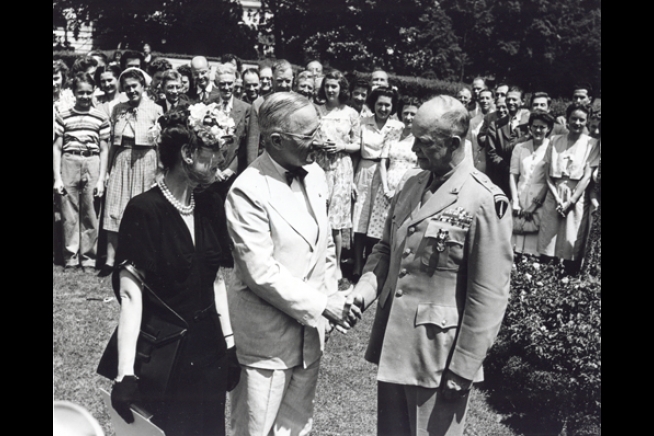 A Photograph of President Harry Truman in grey suit shaking hands with Dwight Eisenhower in a general's uniform in 1945. Mamie Eisenhower, dressed in a black dress, stands next to Dwight Eisenhower