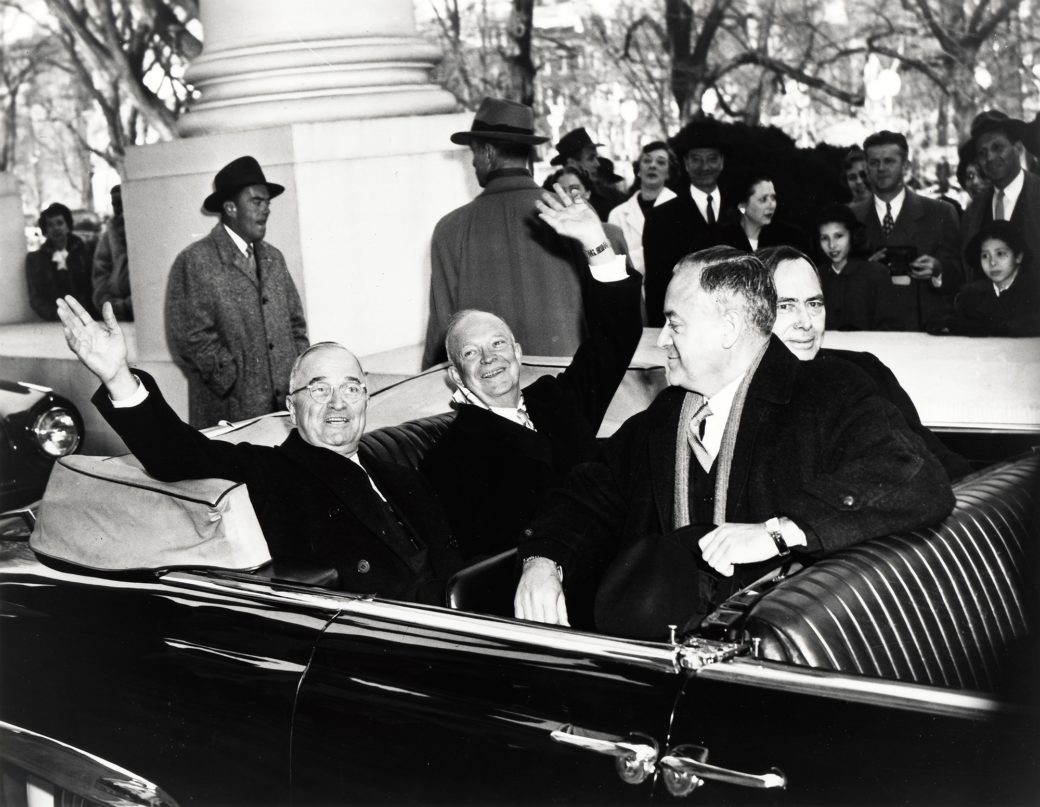 Black and White Photograph of Harry Truman and Dwight Eisenhower , both smiling, both wearing black suits and coats, in the back of a convertible black vehicle. A number of people stand in the background and two other men are seated in front. 