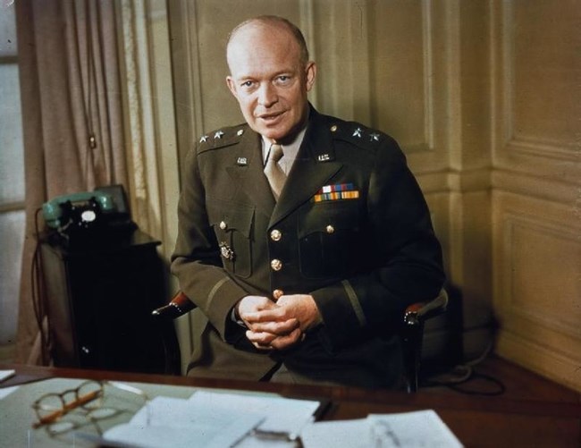 Dwight Eisenhower, wearing a dark green military uniform, with two stars on shoulders, tan shirt and tan tie, sits with his hands folded across his lap. In front of him is a large brown table, upon which are papers and Eisenhower's pair of glasses.