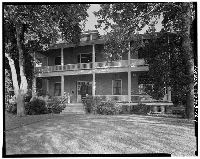 Black and White Photograph of a large, two-story brick building with wrap around porch and several large columns stands at Fort Sam Houston in Texas. In front of the building are two large trees.