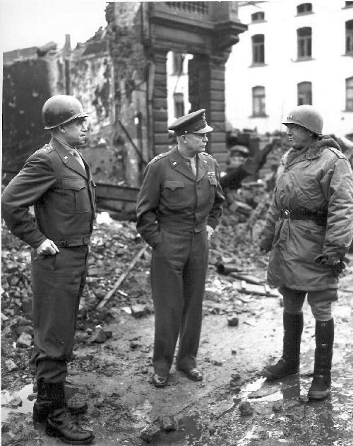 Three generals of the United States army stand in front of a destroyed building in Bastogne on a winter day