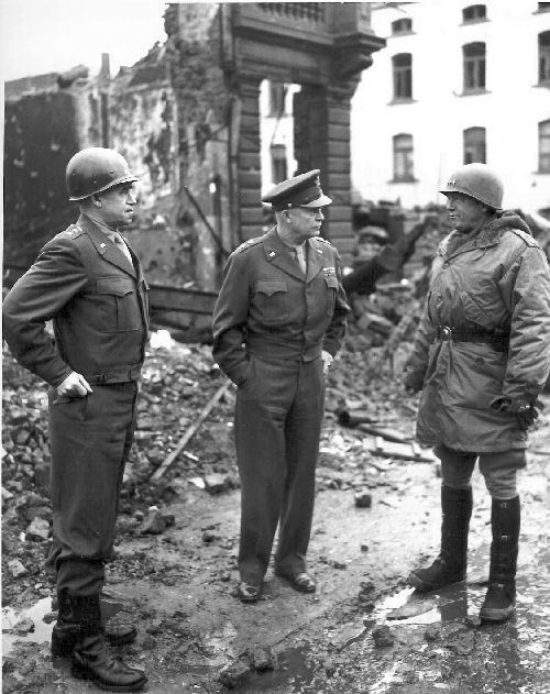 Three generals of the United States army stand in front of a destroyed building in Bastogne on a winter day