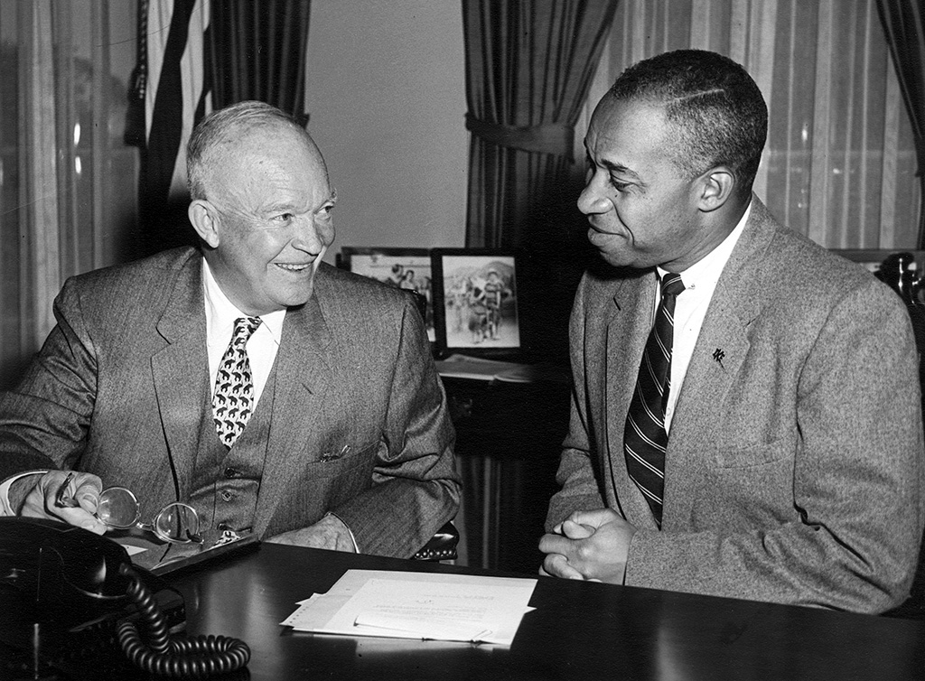 President Eisenhower and E. Frederic Morrow, both wearing gray suits, are seated behind a large desk. Eisenhower is holding his glasses and both men are smiling. A black rotary phone is on the desk.